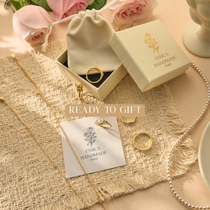 May include: An assortment of gold-colored jewelry, including rings and a necklace, is displayed on a textured beige surface. A small box and a drawstring pouch hold some of the jewelry. The text "READY TO GIFT" is visible, along with the brand name "ESSICA HANDMADE PARIS".
