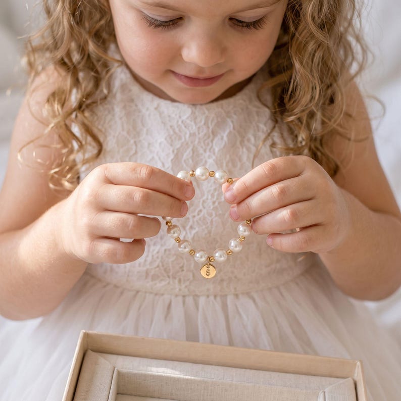 May include: A beaded bracelet featuring alternating white pearls and small gold-toned metallic spheres, finished with a circular gold-toned charm engraved with the letter S. The bracelet is held in place, showcasing the contrast between the white beads.