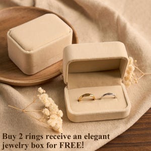 May include: Two rings, one gold and one silver, displayed in an open beige jewelry box. The box is on a beige cloth surface, with a closed matching box and a wooden plate in the background. The text at the bottom says "Buy 2 rings receive an elegant jewelry box for FREE!"