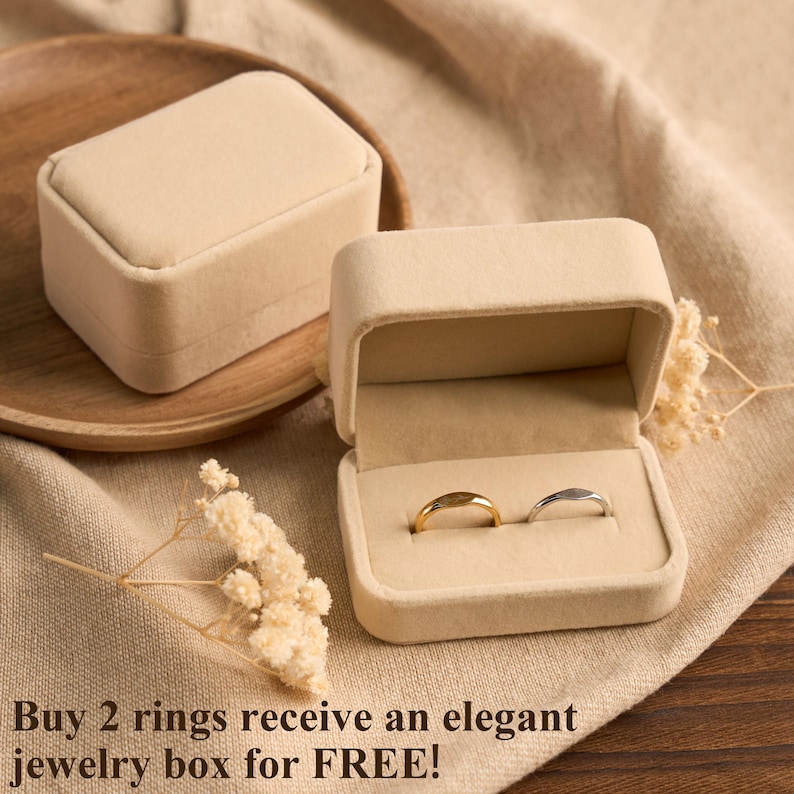 May include: Two rings, one gold and one silver, displayed in an open beige jewelry box. The box is on a beige cloth surface, with a closed matching box and a wooden plate in the background. The text at the bottom says "Buy 2 rings receive an elegant jewelry box for FREE!"