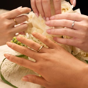 May include: Close-up of three hands wearing rings. Two gold rings with a small diamond are visible, along with a silver ring. The rings are on fingers in front of a bouquet of white flowers.