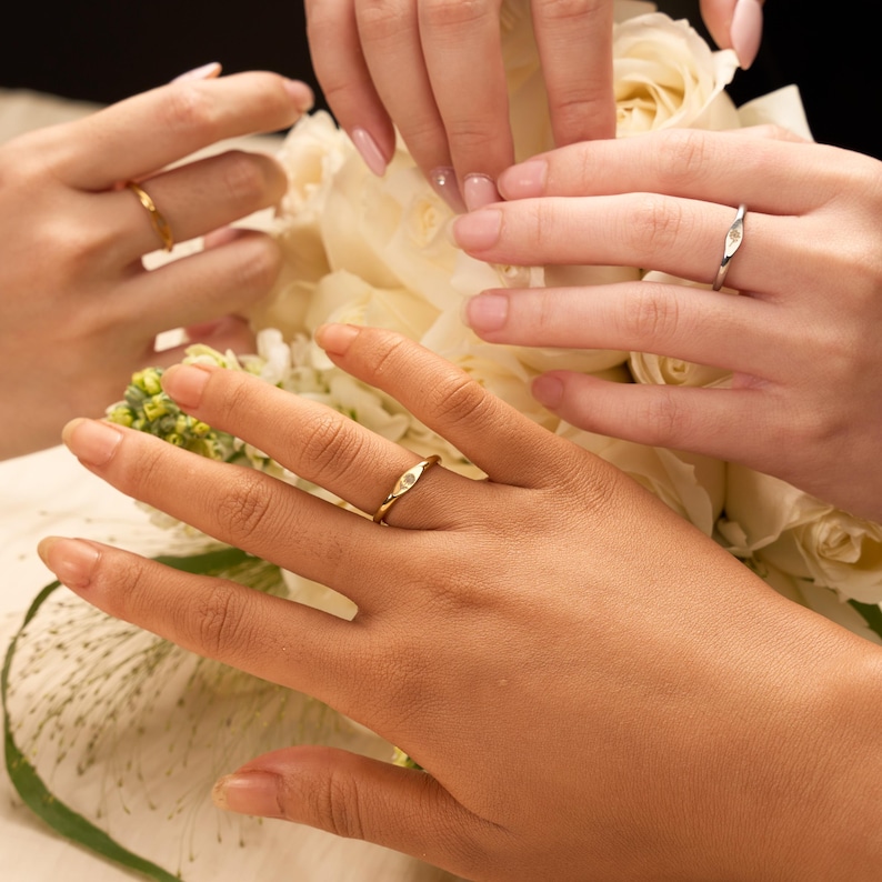 May include: Close-up of three hands wearing rings. Two gold rings with a small diamond are visible, along with a silver ring. The rings are on fingers in front of a bouquet of white flowers.