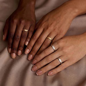 以下が含まれることがあります： さまざまな肌の色合いの手元に、楕円形のデザインの3つの指輪が写っています。2つの指輪は金色で、1つは銀色です。指輪はニュートラルカラーの背景にあります。