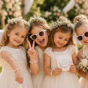 May include: Four children wearing white lace dresses and baby's breath flower crowns. They wear pearl bracelets with gold initial charms. One child wears white sunglasses, another wears pink sunglasses, and one holds a card that says Thank you for being our flower girl.