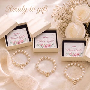 May include: Three white gift boxes containing cards that read "Emma, Will you be my Flower Girl?", "Sophie, Will you be my Flower Girl?", and "Mia, Will you be my Flower Girl?" with three matching pearl bead bracelets featuring gold initial charms.