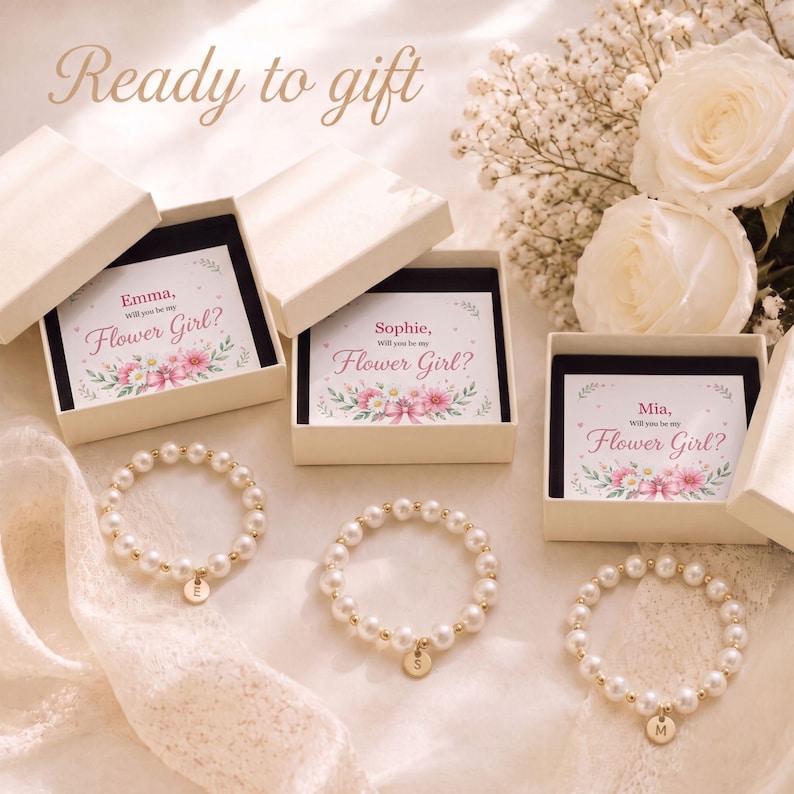 May include: Three white gift boxes containing cards that read "Emma, Will you be my Flower Girl?", "Sophie, Will you be my Flower Girl?", and "Mia, Will you be my Flower Girl?" with three matching pearl bead bracelets featuring gold initial charms.