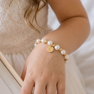 May include: A beaded bracelet featuring alternating white faux pearls and small gold-toned metallic spheres. A circular gold-toned charm hangs from the center of the bracelet, engraved with the capital letter E in a serif font style.