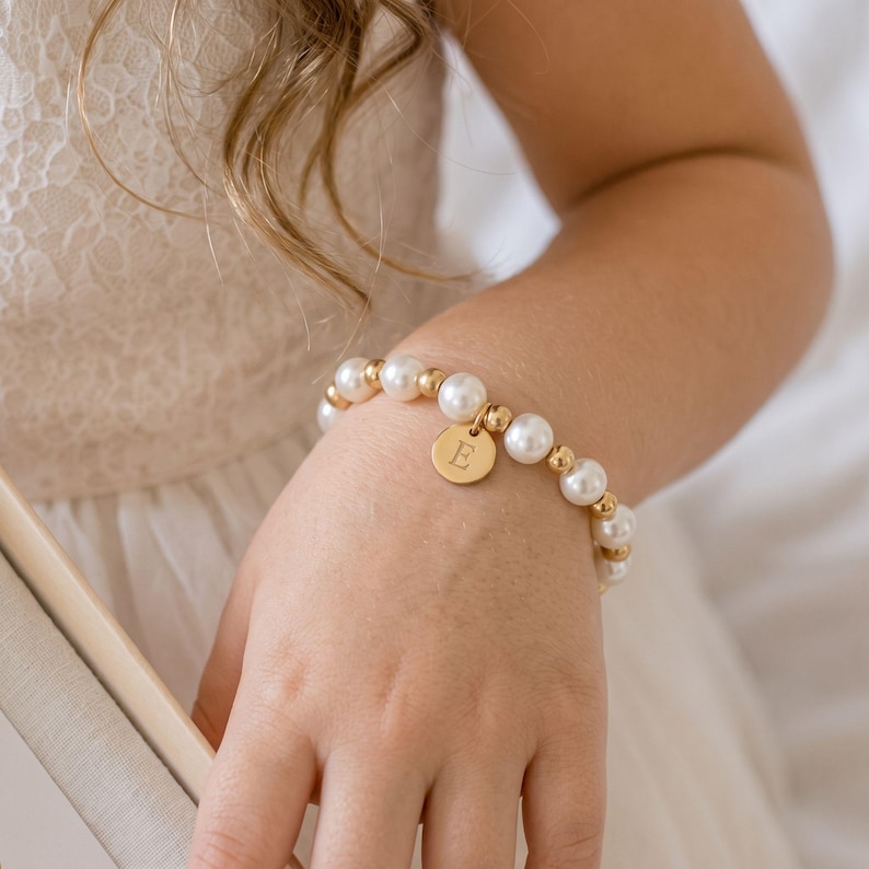 May include: A beaded bracelet featuring alternating white faux pearls and small gold-toned metallic spheres. A circular gold-toned charm hangs from the center of the bracelet, engraved with the capital letter E in a serif font style.