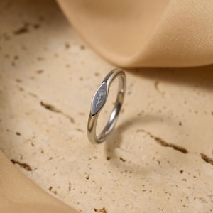 May include: A silver ring with a floral design engraved on an oval-shaped face. The ring is set against a neutral background, highlighting its polished metal finish and delicate detailing. The ring's design is simple and elegant.