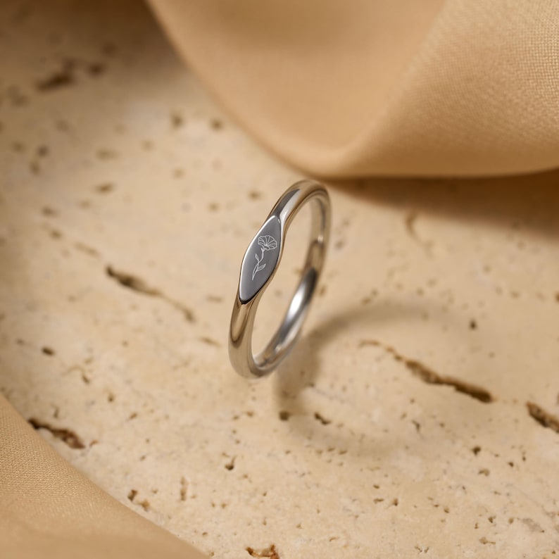 May include: A silver ring with a floral design engraved on an oval-shaped face. The ring is set against a neutral background, highlighting its polished metal finish and delicate detailing. The ring's design is simple and elegant.