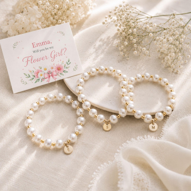 May include: Three beaded bracelets made of white faux pearls and small gold-toned metal spacers. Each bracelet features a small, round gold-toned charm engraved with a single letter: E, S, or M. A white card reads: Emma, Will you be my Flower Girl?