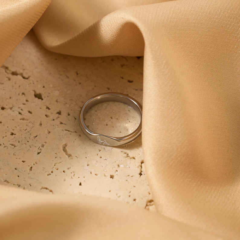 May include: A silver ring with a subtle engraved design. The ring is set against a textured, light-colored surface and a draped, peach-colored fabric. The ring's design is a delicate, minimalist style.