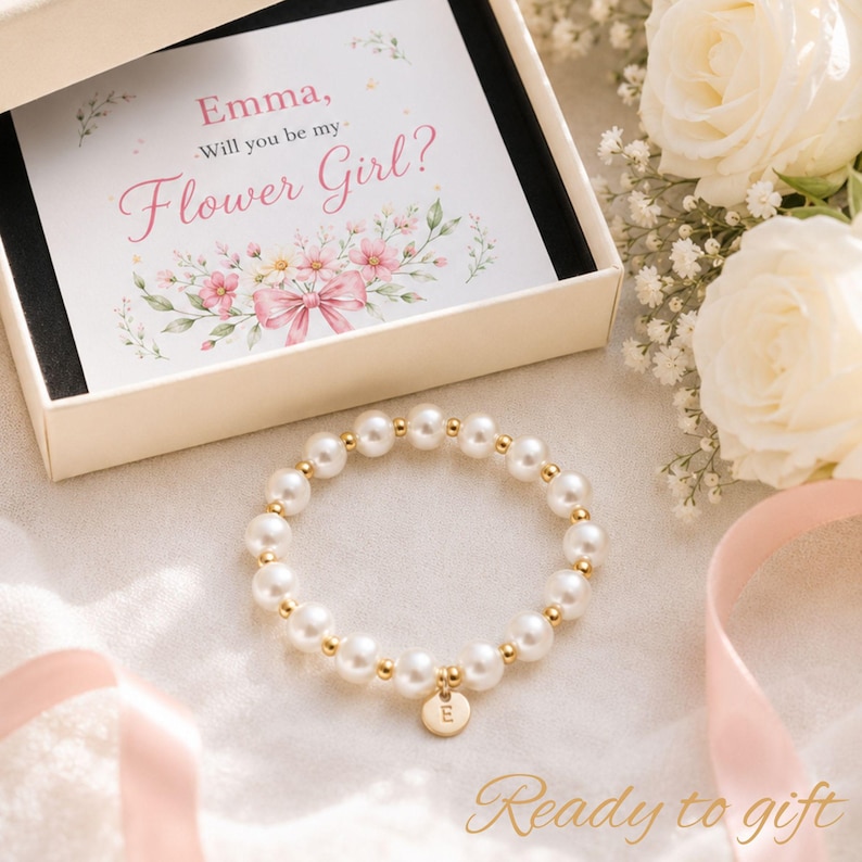 May include: A white pearl bead bracelet with small gold-tone metal spacers and a round gold-tone charm featuring the letter E. The bracelet is presented in an open cream-colored box with a card that reads, Emma, Will you be my Flower Girl?