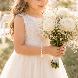 May include: A white pearl bracelet featuring a small gold-toned circular charm engraved with the letter E. The bracelet is worn on a wrist alongside a bouquet of white roses and baby's breath, paired with a sleeveless white lace dress.
