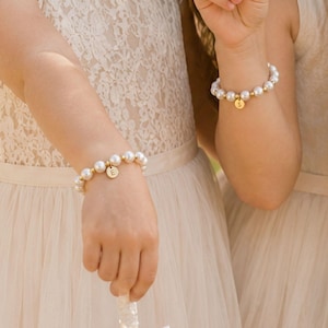 May include: Two beaded bracelets featuring white spherical beads interspersed with small gold-toned metallic spheres. Each bracelet includes a circular gold-toned charm, one engraved with the letter E and the other engraved with the letter S.