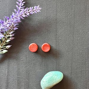 Puede incluir: Dos pequeños pendientes de botón redondos de color coral se muestran sobre una superficie gris texturizada. También hay una ramita de flores de color lavanda y una pequeña piedra ovalada de color verde claro.