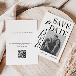 Könnte beinhalten: Zwei Hochzeitseinladungskarten auf einer Holzoberfläche. Eine Karte trägt die Aufschrift "Formal Invitation to Follow" und enthält einen QR-Code. Die andere Karte trägt die Aufschrift "Save the Date" mit einem Foto eines Paares und dem Datum "23 04 26".