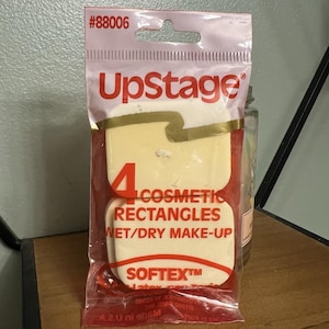 May include: A package of UpStage cosmetic rectangles for wet/dry make-up. The package is pink and red with the number 88006 at the top. The package contains four beige cosmetic rectangles. The brand name is UpStage.