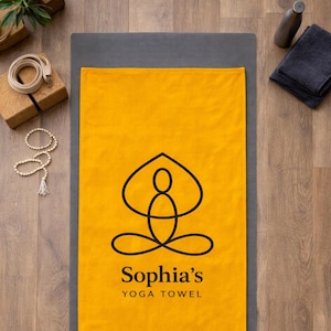 May include: Mustard yellow yoga towel with a black yoga pose graphic and the text "Sophia's YOGA TOWEL". The towel is laid on a gray yoga mat. Other yoga accessories are in the background, including blocks, a water bottle, and candles.