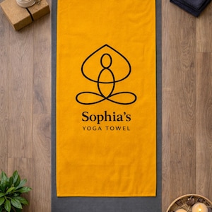 May include: A rectangular mustard yellow yoga towel featuring a black line art graphic of a person in a seated meditation pose. Below the graphic, the text reads Sophia's YOGA TOWEL in black, sans-serif and serif fonts, centered on the towel.