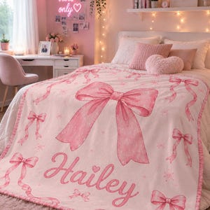 May include: A pink and white blanket with a large pink bow design and the name "Hailey" in cursive script. The blanket features multiple smaller bows and ribbon accents. The bedding is on a bed with pink pillows and a heart-shaped pillow.