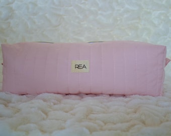 Può includere: Un beauty case rettangolare trapuntato rosa chiaro con cuciture verticali. La borsa presenta una piccola etichetta rettangolare con le lettere "REA" in nero. La borsa è appoggiata su una superficie bianca strutturata.