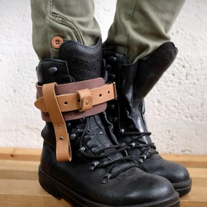Puede incluir: Botas de combate de cuero negro con un detalle de correa de cuero marrón. Las botas tienen un diseño robusto con cordones y una suela gruesa. Las botas se usan con pantalones verde oliva.