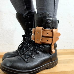 Puede incluir: Botas de combate de cuero negro con cordones y una correa de tobillo de cuero marrón con hebillas. Las botas se usan con vaqueros grises. Las botas están sobre una superficie de madera.