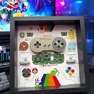 Könnte beinhalten: Ein schwarzer Schattenfugenrahmen mit einem zerlegten Super Famicom-Controller. Das Display zeigt das Controller-Gehäuse, eine grüne Hauptplatine, Gummipads, Trigger und beschriftete Tasten, angeordnet auf einem farbenfrohen Hintergrund im Nintendo-Design.