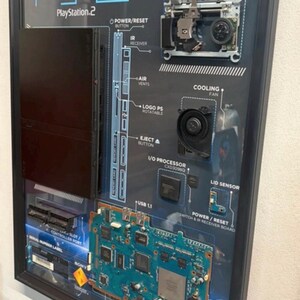 May include: A framed wall display featuring a disassembled black PlayStation 2 console. The components, including the optical disc drive, cooling fan, motherboard, and various internal parts, are mounted on a dark background with white labels.