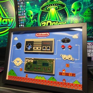 May include: Framed Nintendo controller art piece with a blue background. Features a controller, circuit board, and Mario Bros. scene. Includes the Nintendo logo and text. The frame is black.