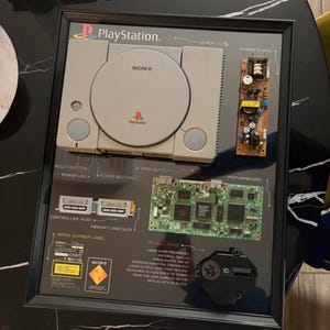 May include: A framed deconstructed PlayStation console display featuring the gray console shell, a power board, a motherboard, and an optical drive. The display includes labels for components like the reset button, controller port, and laser diode properties.