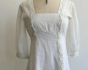 May include: A floor-length white dress featuring a square neckline, three-quarter length sheer sleeves, and vertical floral lace trim detailing that runs from the shoulders down to the hemline. The garment has a fitted bodice and a long skirt.