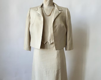 May include: A beige skirt suit with a matching jacket and skirt. The jacket has a notched collar and three-quarter sleeves. The skirt is knee-length and has a straight silhouette.