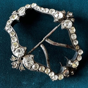 May include: An antique silver-toned buckle, encrusted with numerous clear and yellow-toned gemstones. The buckle has a decorative, openwork design with a central pin and a curved, shield-like shape. The gemstones are set closely together, creating a sparkling effect.