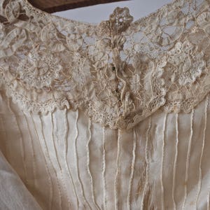 May include: A close-up of the back of a white cotton blouse with a lace collar. The collar is made of delicate white lace with a floral pattern. The blouse has a button closure at the back of the neck.