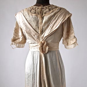 May include: A vintage ivory satin dress with a lace neckline and a sash tied at the back. The dress has a fitted bodice and a flowing skirt.