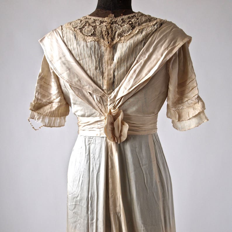 May include: A vintage ivory satin dress with a lace neckline and a sash tied at the back. The dress has a fitted bodice and a flowing skirt.