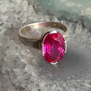 May include: A silver ring featuring a large, oval-cut, vibrant pink gemstone. The ring has a decorative design on the band near the gemstone. The gemstone is the focal point, catching the light and reflecting a bright color.
