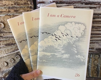 May include: Three identical softcover booklets with cream-colored covers, each featuring the text "I am a Camera" in red at the top and the number "26" at the bottom right. The cover art depicts a flock of birds flying over large, detailed clouds.