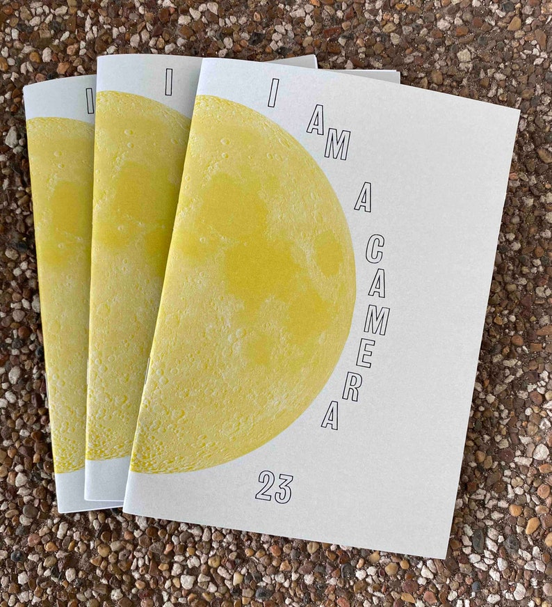 An A5 size zine with a grey cover with an image of a yellow full moon and the title "I am a Camera 23"