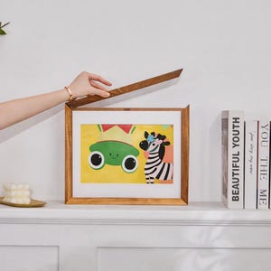 Puede incluir: Un marco rectangular de madera con tapa superior abatible, que muestra una colorida ilustraci&oacute;n de una rana verde de ojos grandes y una cebra de rayas blancas y negras sobre un fondo amarillo. El marco est&aacute; colocado junto a varios libros.
