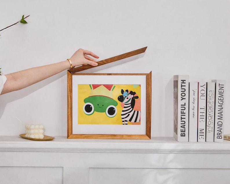 Puede incluir: Un marco rectangular de madera con tapa superior abatible, que muestra una colorida ilustraci&oacute;n de una rana verde de ojos grandes y una cebra de rayas blancas y negras sobre un fondo amarillo. El marco est&aacute; colocado junto a varios libros.
