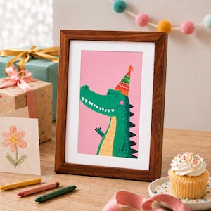 Puede incluir: Un marco de fotos de madera oscura que contiene una impresi&oacute;n de un cocodrilo verde con un sombrero de fiesta a rayas de colores sobre un fondo rosa s&oacute;lido. El cocodrilo tiene una barriga amarilla clara y una amplia sonrisa, en estilo plano.