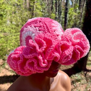 Op de afbeelding: Een handgemaakte gehaakte hoed in roze en wit. De hoed heeft een gerimpelde rand en een getextureerde kroon. Dit unieke accessoire is perfect om een vleugje fantasie aan elke outfit toe te voegen.