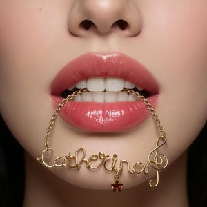 May include: A gold-colored necklace spelling "Catherine" in cursive, with a treble clef and a red star charm. The necklace is held in the mouth, with the focus on the lips and teeth. The lips are glossy and a shade of pink.