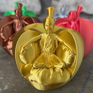 May include: A gold-colored decorative figurine of a seated woman with outstretched arms, atop a heart-shaped base. Other figurines in copper, red, and green are visible in the background. The figurines appear to be 3D printed.