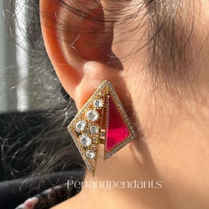 May include: A gold-toned, geometric earring with a diamond shape, featuring a red triangular inlay and clear gemstones. The earring is set with small, clear stones around the edges and has the text "Penandpendant" at the bottom.