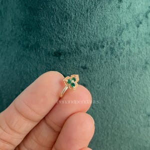 May include: A gold-toned nose ring featuring a floral-shaped setting with three small, round, green stones arranged in a cluster. The metal surface has a textured, decorative finish around the stones. The text Penandpendants is visible.