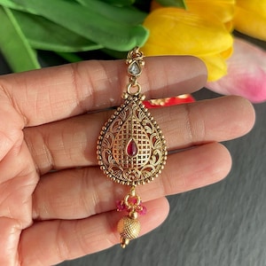 May include: A gold-colored pendant with a teardrop design, featuring a red gemstone at the center and intricate filigree detailing. The pendant is suspended from a delicate chain and adorned with small pink beads and a textured gold ball. The text "Penandpendant" is visible.